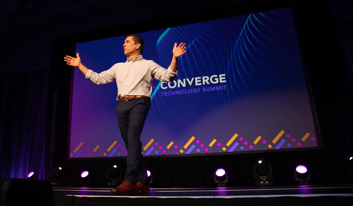 Keynote speaker on stage at the Converge Technology Summit in Milwaukee’s Baird Center, addressing a professional audience under stage lighting.