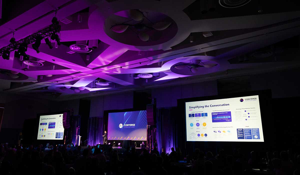 Wide-angle photo of the Converge Technology Summit stage setup in the Baird Center Grand Ballroom in Milwaukee, with lighting, screens, and a speaker platform.